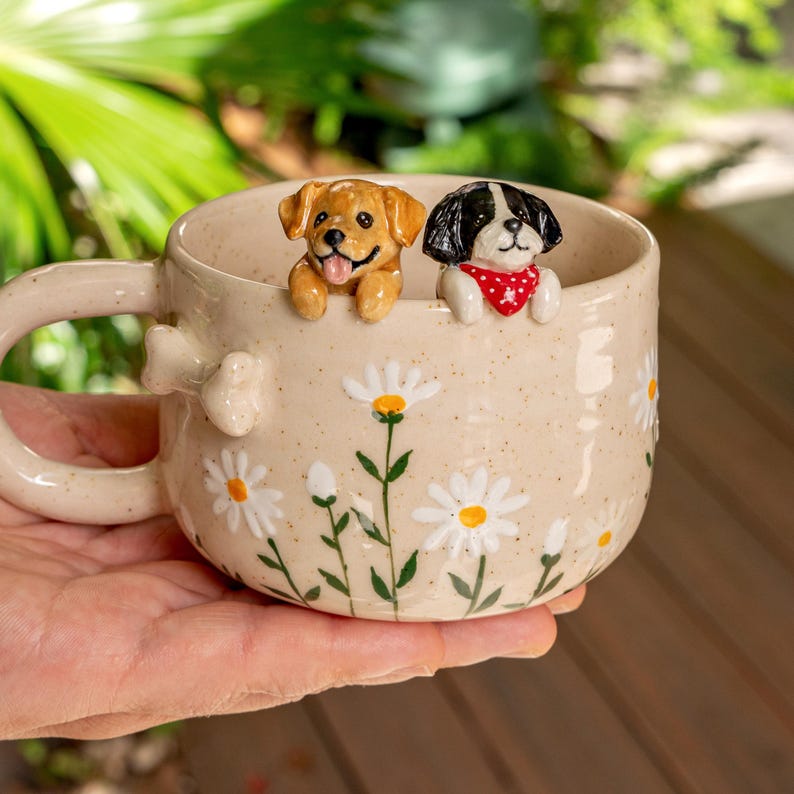 Puede incluir: Una taza de cer&aacute;mica beige con acabado moteado, decorada con margaritas blancas pintadas a mano con centros amarillos. Dos peque&ntilde;as figuras de perros esculpidas est&aacute;n unidas al borde interior y un detalle en forma de hueso est&aacute; en el asa.