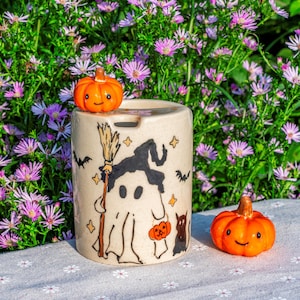 May include: A beige ceramic Halloween-themed container with a ghost, witch hat, broom, bats, and stars design. Two small orange pumpkins with smiling faces sit on the container and beside it. The background features purple flowers.