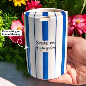 May include: A cylindrical ceramic mug featuring vertical blue and white stripes. The front of the mug displays the handwritten black text "Smoke 'em if you got 'em" centered between the stripes. The mug has a small rectangular cutout at the top rim.