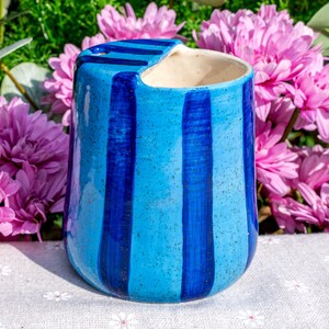 Peut inclure: Un pot en c&eacute;ramique avec un vernis bleu clair mouchet&eacute; et des rayures verticales bleu fonc&eacute;. Le r&eacute;cipient pr&eacute;sente une d&eacute;coupe unique sur le bord sup&eacute;rieur, cr&eacute;ant une ouverture distincte. L'int&eacute;rieur est d'une couleur blanc cass&eacute; lisse.