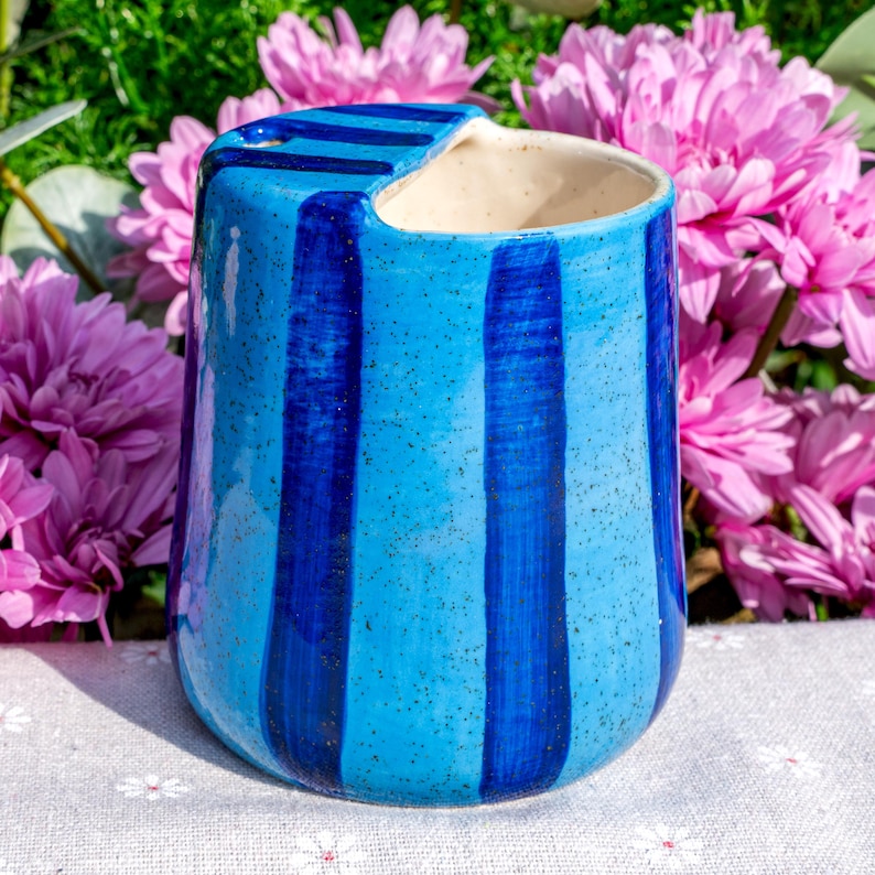 Peut inclure: Un pot en c&eacute;ramique avec un vernis bleu clair mouchet&eacute; et des rayures verticales bleu fonc&eacute;. Le r&eacute;cipient pr&eacute;sente une d&eacute;coupe unique sur le bord sup&eacute;rieur, cr&eacute;ant une ouverture distincte. L'int&eacute;rieur est d'une couleur blanc cass&eacute; lisse.