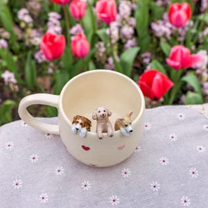 K&ouml;nnte beinhalten: Eine wei&szlig;e Keramiktasse mit drei kleinen Hundefiguren, die &uuml;ber den Rand schauen. Die Tasse hat einen Henkel und rosa Herzen an der Seite. Die Tasse steht auf einer wei&szlig;en Tischdecke mit Blumenmuster.