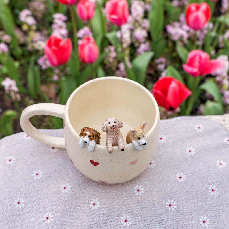 K&ouml;nnte beinhalten: Eine wei&szlig;e Keramiktasse mit drei kleinen Hundefiguren, die &uuml;ber den Rand schauen. Die Tasse hat einen Henkel und rosa Herzen an der Seite. Die Tasse steht auf einer wei&szlig;en Tischdecke mit Blumenmuster.