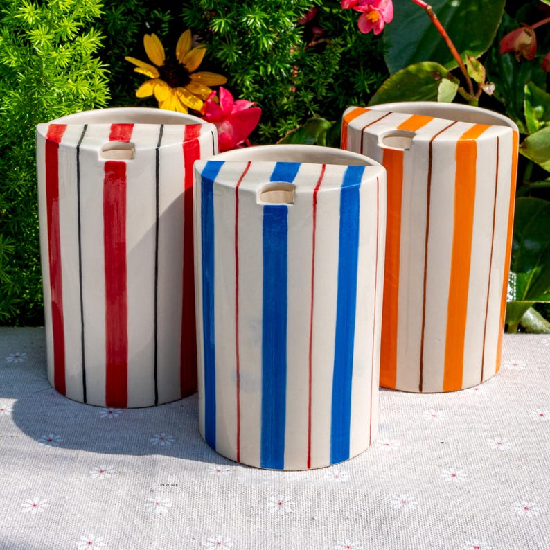 May include: Three cylindrical ceramic containers with flat tops and a single circular opening. Each vessel features a cream-colored base with vertical stripes in red and black, blue and red, or orange and brown, arranged in a repeating pattern.