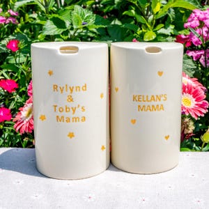 May include: Two off-white ceramic containers with a rectangular shape and a small opening at the top. One reads "Rylind & Toby's Mama" and the other reads "Kellan's Mama" in gold lettering. Gold stars and hearts decorate the containers.