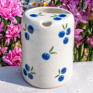 May include: A cylindrical ceramic toothbrush holder with a speckled cream background. The holder is decorated with hand-painted blue blueberries and green leaves. The top has a hole for a toothbrush. The background features pink flowers.