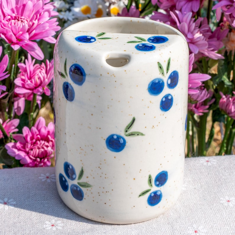 May include: A cylindrical ceramic toothbrush holder with a speckled cream background. The holder is decorated with hand-painted blue blueberries and green leaves. The top has a hole for a toothbrush. The background features pink flowers.