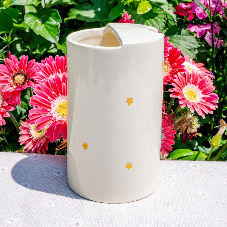 May include: A cream-colored ceramic utensil holder with a handle and yellow star accents. The cylindrical holder is decorated with three small, evenly spaced stars. The background features vibrant pink flowers and green foliage.