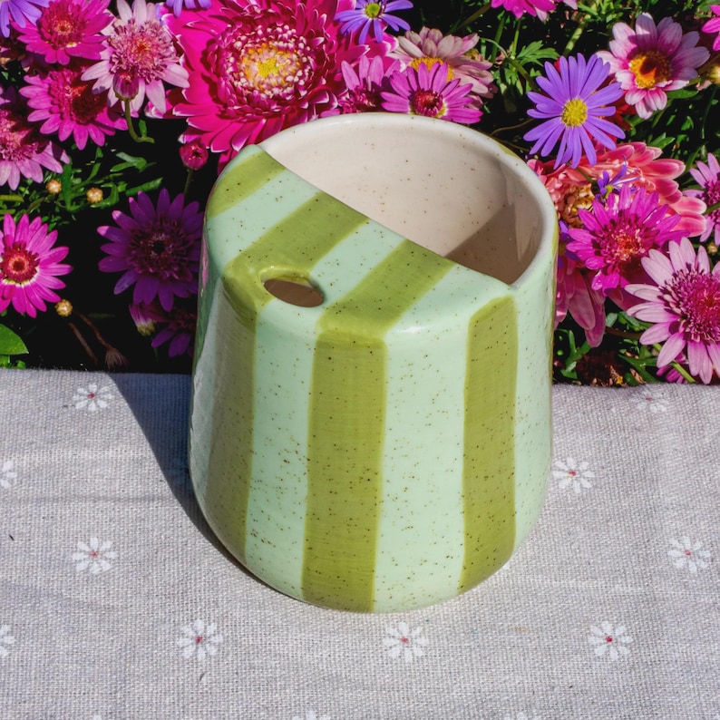 Peut inclure: Une tasse en c&eacute;ramique avec un motif &agrave; rayures verticales vert clair et vert olive mouchet&eacute;. La tasse pr&eacute;sente une forme arrondie lisse avec une petite ouverture circulaire pr&egrave;s du bord sup&eacute;rieur, un int&eacute;rieur cr&egrave;me et une finition brillante.