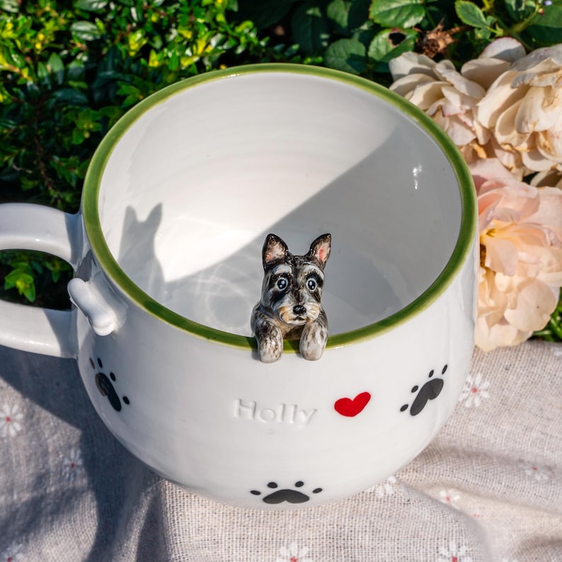 May include: A white ceramic mug with a green rim featuring a small, hand-painted three-dimensional schnauzer dog figure perched on the inner edge. The exterior is decorated with black paw prints, a red heart, and the embossed name Holly.