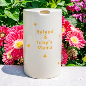 May include: A cream-colored ceramic tumbler with the text "Rylind & Toby's Mama" in gold lettering, accented with gold star designs. The tumbler is cylindrical with a small opening at the top. The background features vibrant pink and red flowers.