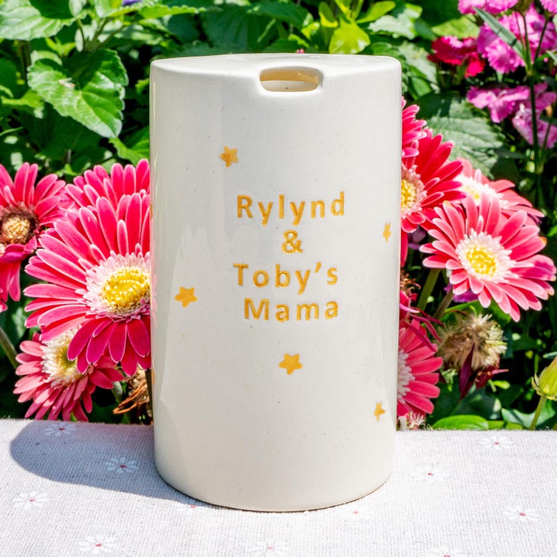May include: A cream-colored ceramic tumbler with the text "Rylind & Toby's Mama" in gold lettering, accented with gold star designs. The tumbler is cylindrical with a small opening at the top. The background features vibrant pink and red flowers.