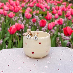 K&ouml;nnte beinhalten: Eine wei&szlig;e Keramiktasse mit rosa Herzen und drei kleinen Hundefiguren, die &uuml;ber den Rand schauen. Die Tasse steht auf einer wei&szlig;en Tischdecke mit Blumenmuster.