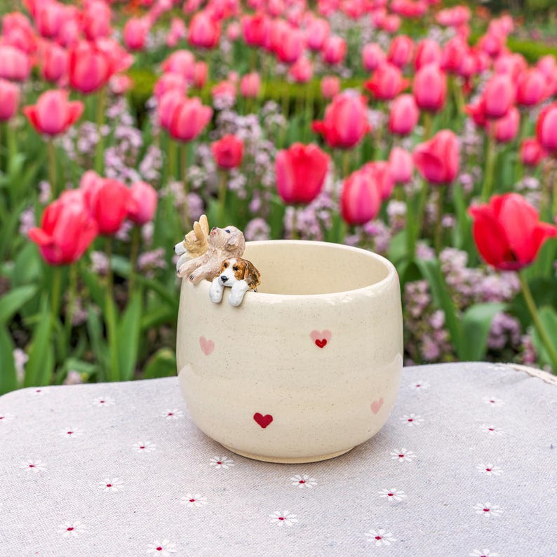 K&ouml;nnte beinhalten: Eine wei&szlig;e Keramiktasse mit rosa Herzen, die auf die Seite gedruckt sind. Drei kleine Hundefiguren schauen &uuml;ber den Rand der Tasse.