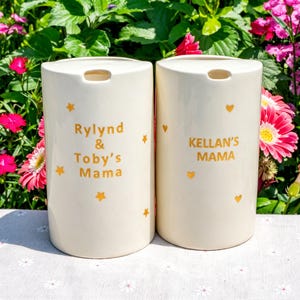 May include: Two off-white ceramic vases with a cylindrical shape. One vase has the text "Rylind & Toby's Mama" and the other reads "Kellan's Mama" in gold lettering. Gold stars and hearts decorate the vases.