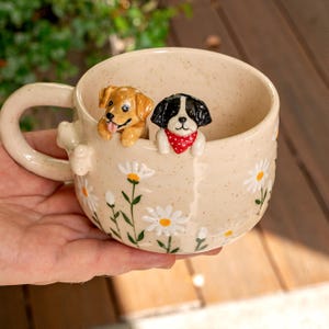 Puede incluir: Una taza de cer&aacute;mica color crema con acabado moteado y un patr&oacute;n de margaritas pintadas a mano. Dos peque&ntilde;as figuras de perros esculpidas est&aacute;n unidas al borde interior: un cachorro marr&oacute;n dorado y un cachorro blanco y negro con pa&ntilde;uelo rojo.