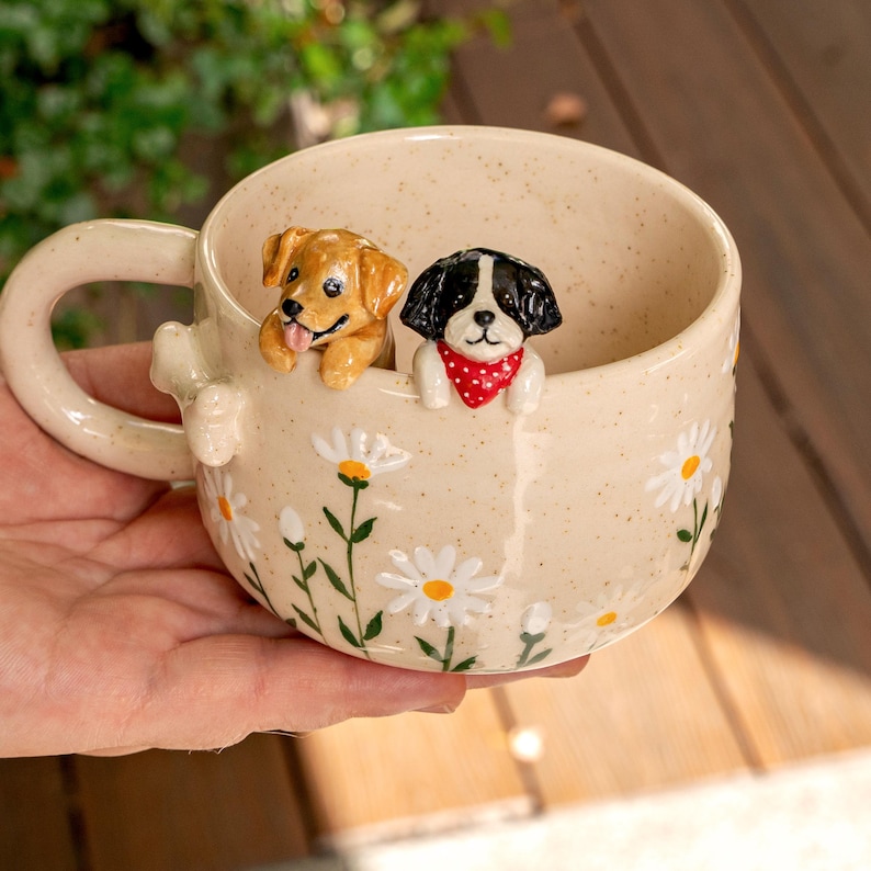 Puede incluir: Una taza de cer&aacute;mica color crema con acabado moteado y un patr&oacute;n de margaritas pintadas a mano. Dos peque&ntilde;as figuras de perros esculpidas est&aacute;n unidas al borde interior: un cachorro marr&oacute;n dorado y un cachorro blanco y negro con pa&ntilde;uelo rojo.