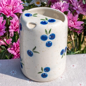 May include: A cylindrical ceramic utensil holder with a speckled cream-colored finish. The holder is decorated with hand-painted blue blueberries and green leaves. The holder has a curved top with a small hole. The background features pink flowers.