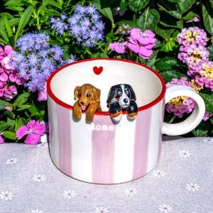 May include: A striped ceramic mug in pink and white, with two dog figurines perched on the rim. The mug has a red inner rim and a small red heart. The word "Honey" is written on the side of the mug.