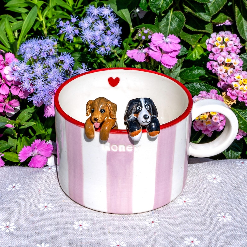 May include: A striped ceramic mug in pink and white, with two dog figurines perched on the rim. The mug has a red inner rim and a small red heart. The word "Honey" is written on the side of the mug.