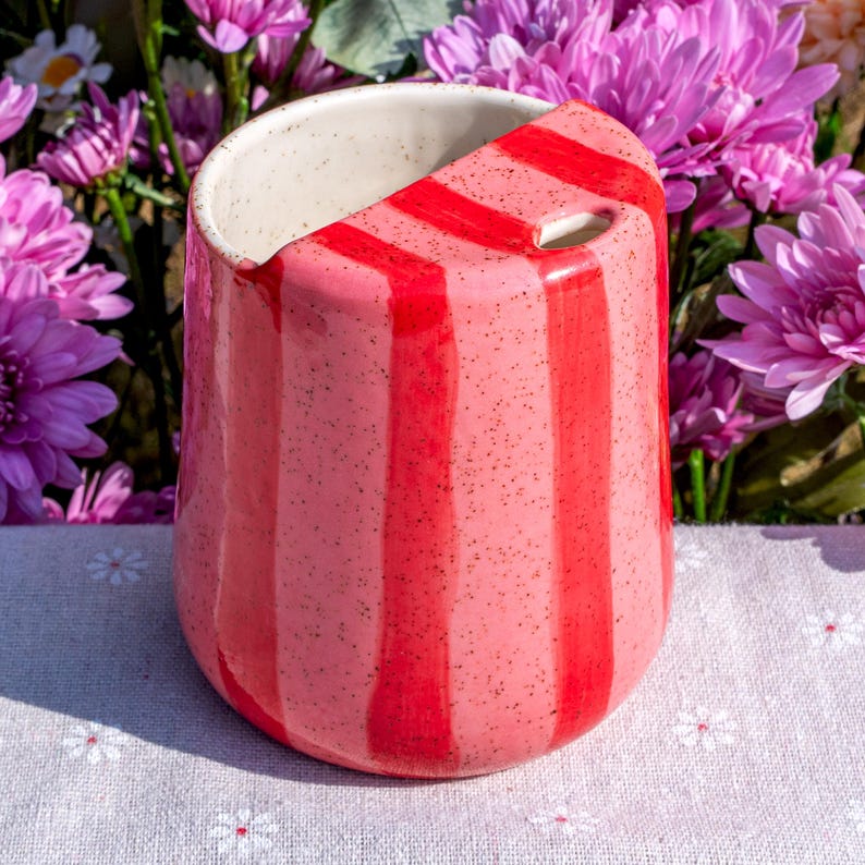 Peut inclure: Un gobelet en c&eacute;ramique avec une base rose tachet&eacute;e et des rayures verticales rouge vif. Le r&eacute;cipient pr&eacute;sente un bord inclin&eacute; unique qui cr&eacute;e un couvercle partiel sur l'ouverture sup&eacute;rieure, laissant un petit trou circulaire pour boire.