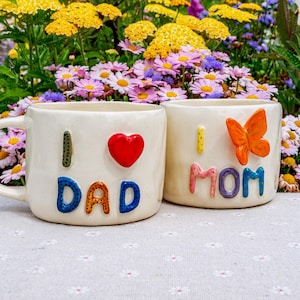 May include: Two off-white ceramic mugs with the words "I LOVE DAD" and "I MOM" in colorful, raised lettering. One mug has a red heart, the other an orange butterfly. The mugs are set against a backdrop of colorful flowers.