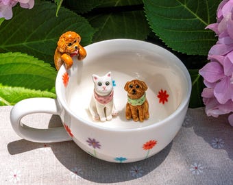 May include: A white ceramic mug decorated with small red, purple, and blue flower patterns. Inside the mug are two small animal figurines, a white cat and a brown dog, while a second brown dog figurine is perched on the rim of the mug.