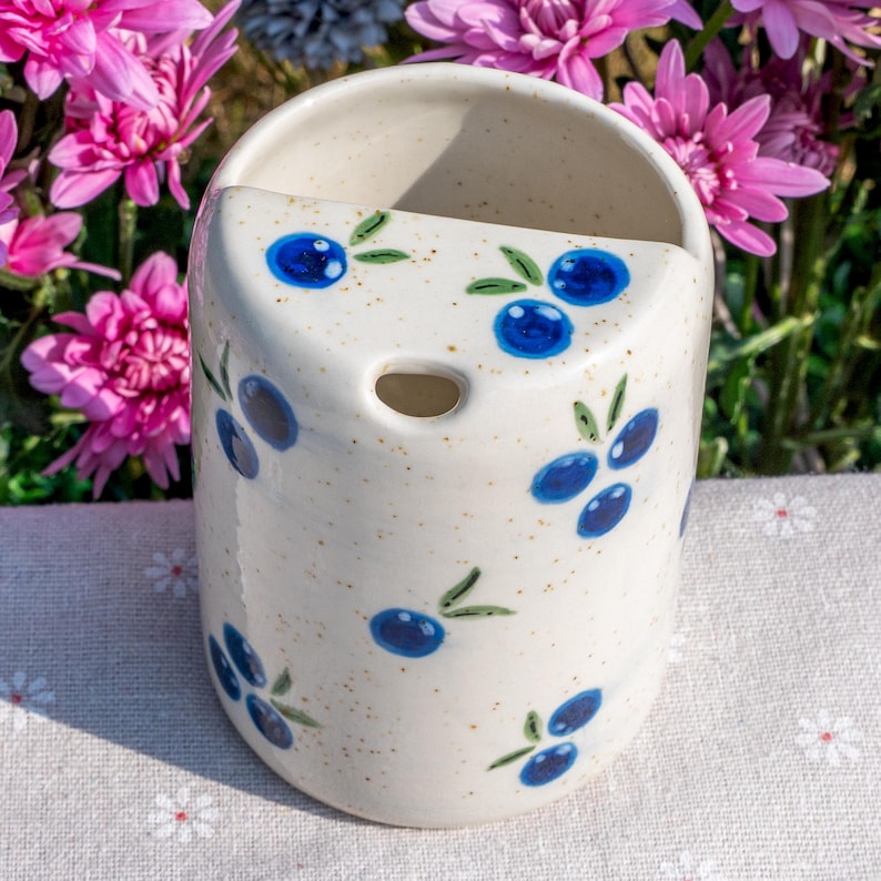 May include: A cream-colored ceramic wall pocket with a hand-painted blueberry design. The cylindrical pocket has a curved top and a hole near the top. The blueberries are blue with green leaves. The background includes pink flowers.