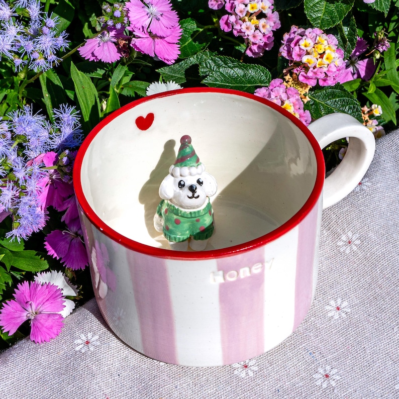 May include: A ceramic mug with pink and white stripes, a red rim, and a white handle. Inside, a small figurine of a white dog wearing a green sweater and hat. A red heart is painted inside the rim. The word "Honey" is written on the side.