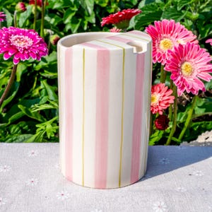 May include: A cylindrical ceramic utensil holder featuring a cream-colored base with vertical stripes in soft pink and thin olive green lines. The top edge has a notched cutout section, and the surface has a smooth, glossy finish throughout.