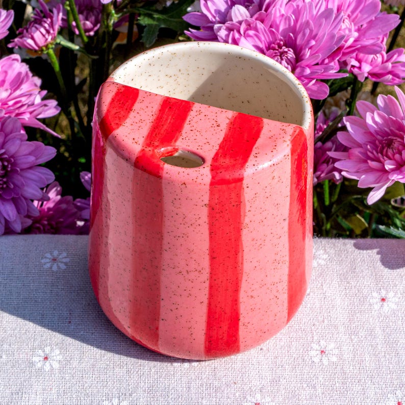 Peut inclure: Un pot de fleurs mural en c&eacute;ramique avec une ouverture semi-circulaire sur le dessus et un petit trou circulaire pour la suspension. L'ext&eacute;rieur est orn&eacute; de rayures verticales alternant des tons rose clair et rouge vif, avec une texture mouchet&eacute;e.
