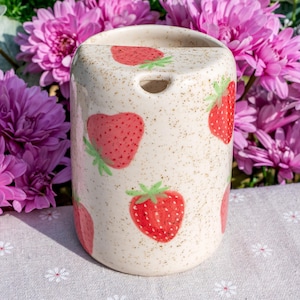 Könnte beinhalten: Ein zylindrischer Zahnbürstenhalter aus Keramik mit gesprenkelter, cremefarbener Oberfläche. Der Halter ist mit roten Erdbeeren und grünen Blättern verziert. Ein kleines Loch ist oben zu sehen. Der Hintergrund zeigt rosa Blüten.