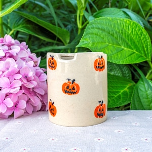 May include: A cylindrical, off-white ceramic container decorated with orange jack-o'-lantern faces. The container has a rectangular opening at the top. The background features pink flowers and green foliage.