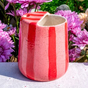 Peut inclure: Un gobelet en c&eacute;ramique avec un &eacute;mail rose clair mouchet&eacute; et des rayures verticales rouge vif. Le r&eacute;cipient pr&eacute;sente un design de bord inclin&eacute; unique, cr&eacute;ant une ouverture asym&eacute;trique au sommet. L'int&eacute;rieur est une finition en c&eacute;ramique blanc cass&eacute;.