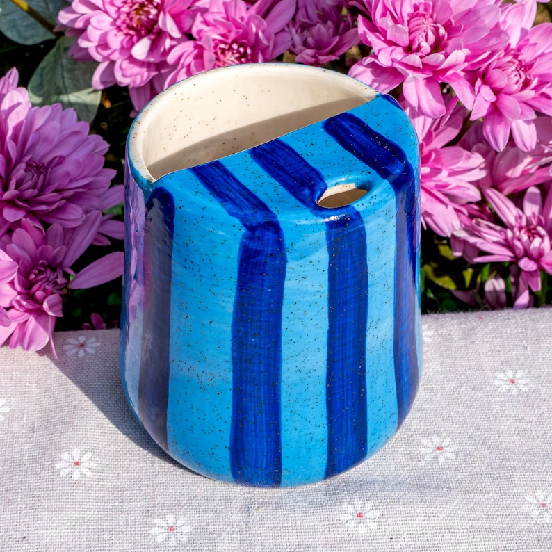 Peut inclure: Une tasse en c&eacute;ramique avec un &eacute;mail bleu clair mouchet&eacute; et des rayures verticales bleu fonc&eacute;. Le r&eacute;cipient pr&eacute;sente un bord sup&eacute;rieur inclin&eacute; et une petite ouverture circulaire pr&egrave;s du rebord. L'int&eacute;rieur est blanc cass&eacute; mouchet&eacute; et lisse.