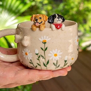Puede incluir: Una taza de cer&aacute;mica beige moteada con margaritas blancas pintadas a mano y tallos verdes. Dos peque&ntilde;as figuras de perros esculpidas, una dorada y otra blanca y negra con un pa&ntilde;uelo rojo, asoman por el borde. Un peque&ntilde;o hueso est&aacute; en el asa.