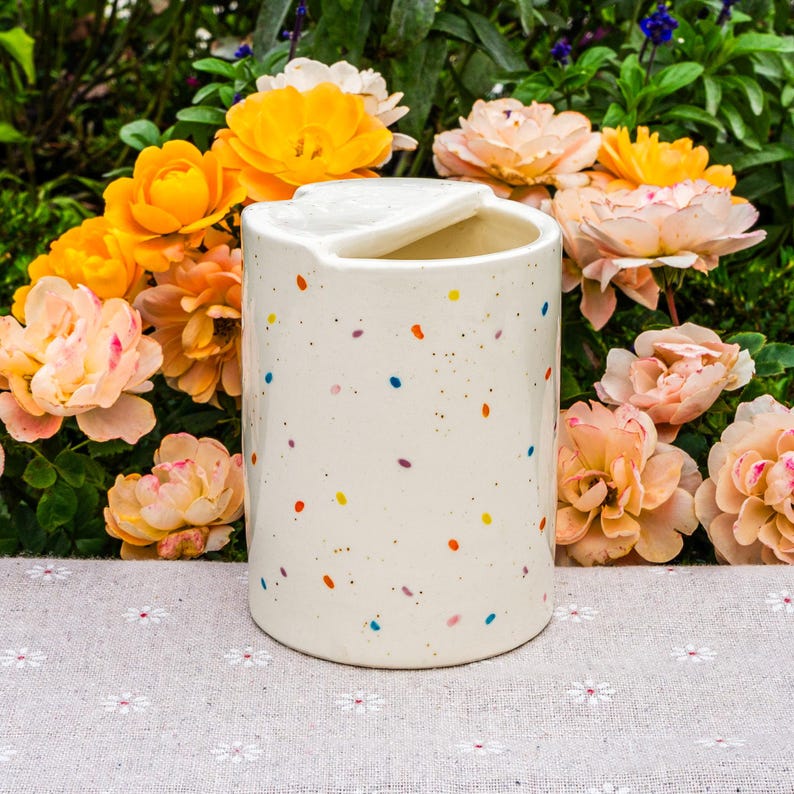 Pode incluir: Um recipiente de cer&acirc;mica creme com tampa, adornado com bolinhas coloridas. O recipiente cil&iacute;ndrico est&aacute; sobre uma superf&iacute;cie com estampa floral. O fundo apresenta vibrantes flores laranja e rosa.