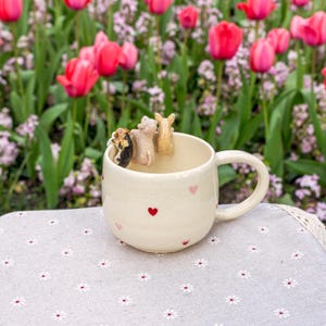 K&ouml;nnte beinhalten: Eine wei&szlig;e Keramiktasse mit rosa Herzen und einer braunen, schwarzen und rosa Figur von drei Tieren im Inneren. Die Tasse steht auf einer wei&szlig;en Tischdecke mit Blumenmuster.