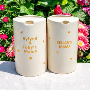 May include: Two off-white ceramic containers with a cylindrical shape. Each container has a small opening at the top and is decorated with gold stars and hearts. One container reads "Rylind & Toby's Mama" and the other reads "Kellan's Mama".