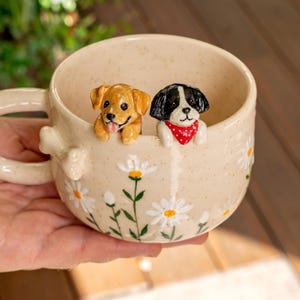 Puede incluir: Una taza de cer&aacute;mica color crema con acabado moteado, decorada con margaritas blancas pintadas a mano con centros amarillos. Dos peque&ntilde;as figuras de perros esculpidas est&aacute;n unidas al borde interior: un cachorro dorado y un cachorro blanco y negro con un pa&ntilde;uelo rojo.