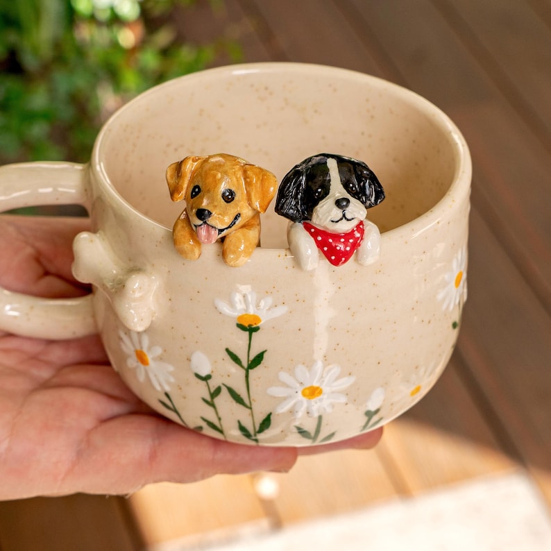 Puede incluir: Una taza de cer&aacute;mica color crema con acabado moteado, decorada con margaritas blancas pintadas a mano con centros amarillos. Dos peque&ntilde;as figuras de perros esculpidas est&aacute;n unidas al borde interior: un cachorro dorado y un cachorro blanco y negro con un pa&ntilde;uelo rojo.