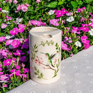 May include: A cream-colored ceramic tumbler with a hummingbird and floral design. The tumbler features a green and pink hummingbird perched on a branch with pink blossoms. The background is a garden of pink and white flowers.