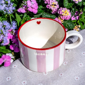 May include: A striped ceramic mug with pink and white vertical stripes. The rim is red, and a small red heart is inside. The word "Honey" is embossed on the side. It has a white handle.