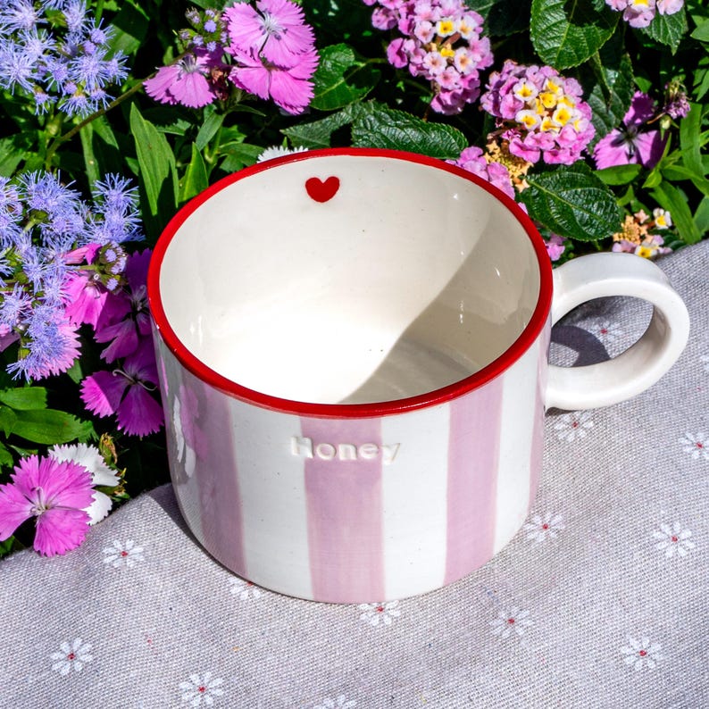 May include: A striped ceramic mug with pink and white vertical stripes. The rim is red, and a small red heart is inside. The word "Honey" is embossed on the side. It has a white handle.
