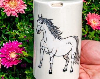 Puede incluir: Un recipiente cilíndrico de cerámica blanca con un dibujo lineal negro detallado de un caballo con crines y cola fluidas. La superficie de la cerámica tiene una textura ligeramente moteada y una pequeña abertura en el borde superior.