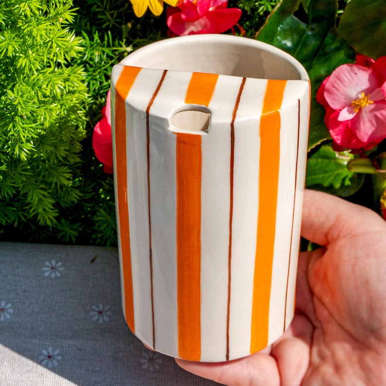 May include: A cylindrical ceramic vessel featuring a cream-colored base with vertical stripes in bright orange and thin brown lines. The top edge has a semi-circular cutout, and the interior is smooth and cream-colored, showing a glossy finish.