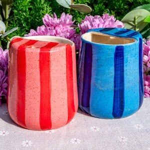 Peut inclure: Deux tasses en c&eacute;ramique avec une finition mouchet&eacute;e. L'une pr&eacute;sente des rayures verticales rouges sur une base rose clair, tandis que l'autre affiche des rayures verticales bleu fonc&eacute; sur une base bleu vif. Les deux ont une forme arrondie.