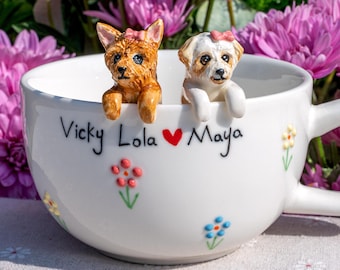 May include: A white ceramic mug featuring hand-painted colorful flowers and the black text Vicky Lola with a red heart and Maya. Two small, sculpted dog figurines, one brown and one white, are perched on the rim, each wearing a pink bow.