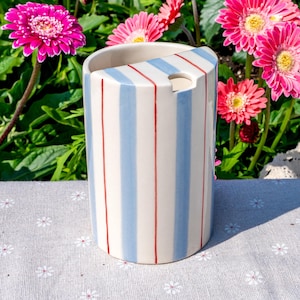 May include: A cylindrical ceramic toothbrush holder featuring a cream-colored base with vertical light blue and thin red stripes. The top edge has a slanted cut-out design with a circular opening for holding items, finished with a smooth glaze.