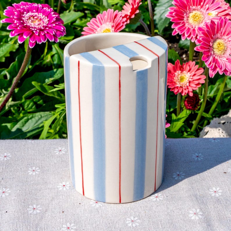 May include: A cylindrical ceramic toothbrush holder featuring a cream-colored base with vertical light blue and thin red stripes. The top edge has a slanted cut-out design with a circular opening for holding items, finished with a smooth glaze.